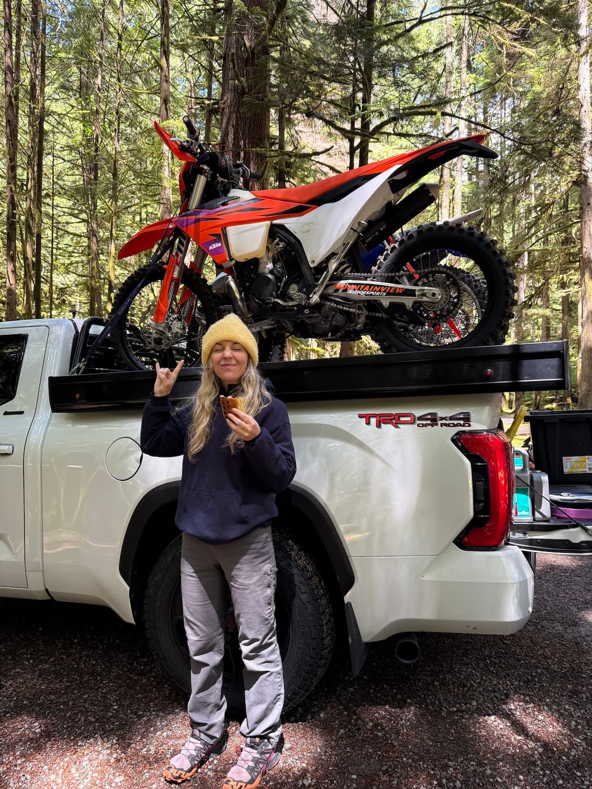Kaiya giving a rock-on sign in front of
her dirt bike, loaded on a truck in the
forest